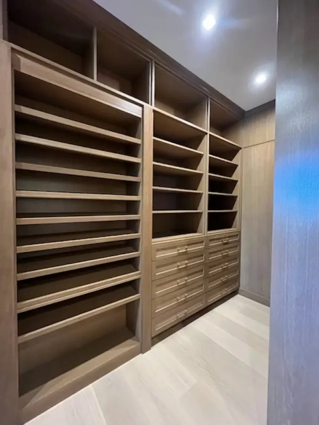 Modern walk-in closet with built-in wooden shelving units and drawers, recessed ceiling lights, and light wood flooring