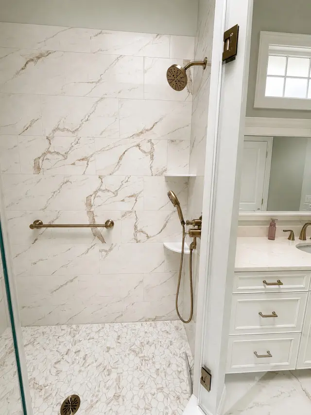 Modern bathroom shower with cream and beige marble tile walls, brass fixtures including a rainfall showerhead and handheld sprayer, grab bar, and textured mosaic tile floor, adjacent to a white vanity with drawers and mirror
