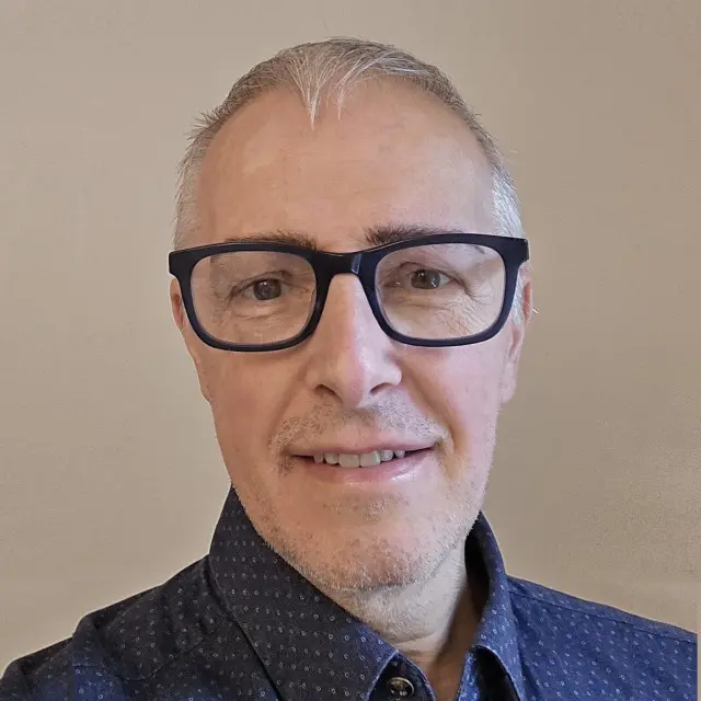Middle-aged person with short gray hair and facial hair wearing black-framed glasses and a dark blue patterned shirt, smiling at the camera against a beige background.
