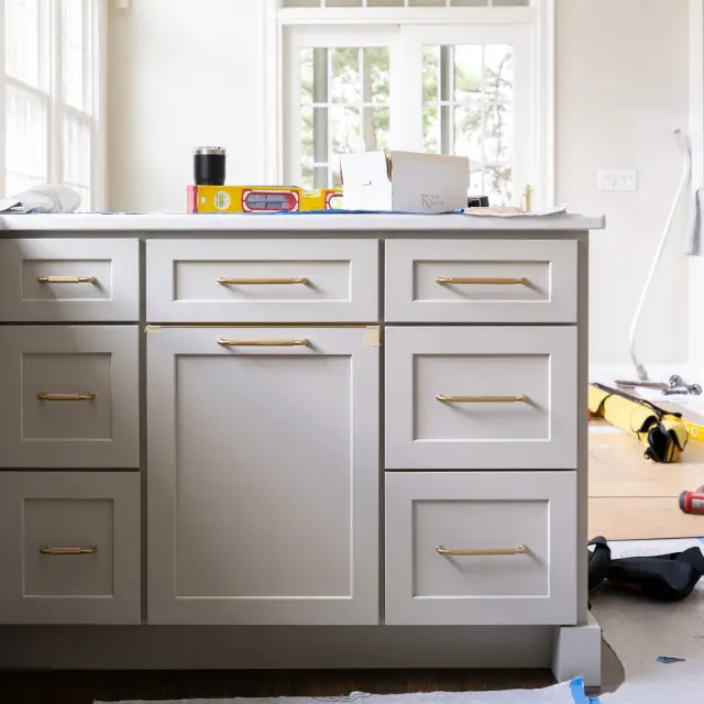 Bright kitchen workspace with white cabinetry and brass handles, countertop displaying construction tools including a yellow level and black thermos, large windows with transom above showing outdoor greenery, hardwood flooring with blue painter's tape, and various tools and materials scattered on floor indicating active renovation or construction project.