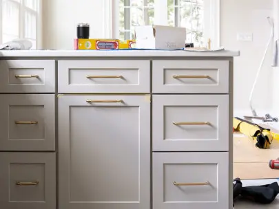 Bright kitchen workspace with white cabinetry and brass handles, countertop displaying construction tools including a yellow level and black thermos, large windows with transom above showing outdoor greenery, hardwood flooring with blue painter's tape, and various tools and materials scattered on floor indicating active renovation or construction project.