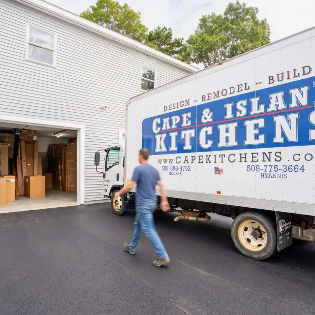 Person in blue shirt running past a white delivery truck with Cape & Island Kitchens branding parked in front of a white residential garage with cardboard boxes visible inside