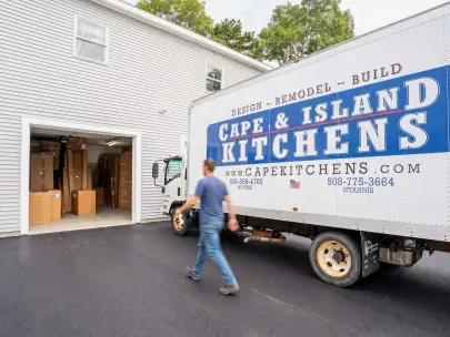Person in blue shirt running past a white delivery truck with Cape & Island Kitchens branding parked in front of a white residential garage with cardboard boxes visible inside