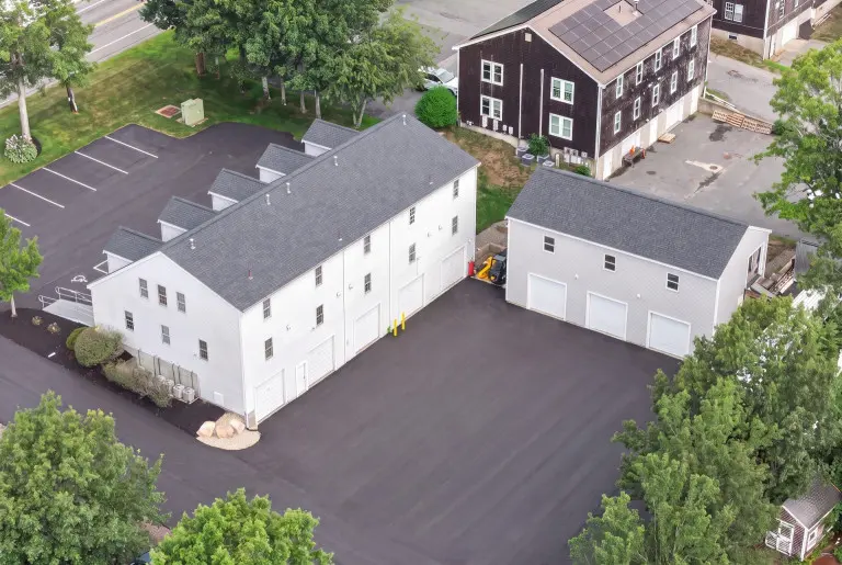 Aerial view of two white residential buildings with gray roofs and multiple garage doors on a paved driveway, surrounded by green trees and landscaping, with a dark brown colonial-style house visible in the background