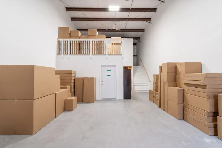 Warehouse interior with cardboard boxes stacked on both sides of a central aisle, a mezzanine level with railing above, white door with green handles, staircase, exposed ceiling beams, and industrial lighting.