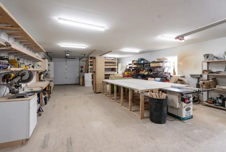Spacious woodworking workshop with fluorescent ceiling lights, white garage door, wooden workbenches and shelving units, power tools on left side, storage containers, lumber and materials organized throughout, and concrete floor.