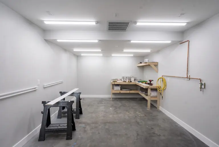 Bright workshop or garage interior with white walls and fluorescent ceiling lights, featuring a workbench with wooden shelving on the right, a sawhorse with white board on the left, concrete floor, coiled yellow hose, copper piping on the wall, and various tools and supplies organized throughout the space.