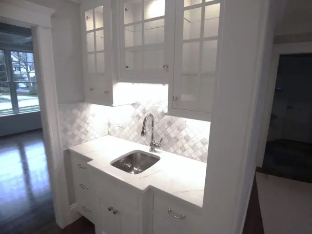 Modern white kitchen with marble countertop, undermount sink, chrome faucet, and marble chevron backsplash, featuring tall windows above and white cabinetry below